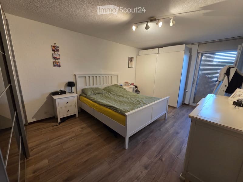 Cozy bedroom with wooden flooring, white bed frame, green bedding, nightstand with lamp, and a small table with a camera on it.