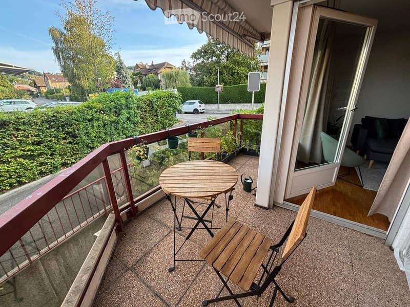 Balcony with a small round table and two wooden chairs, overlooking a residential area with greenery and parked cars.