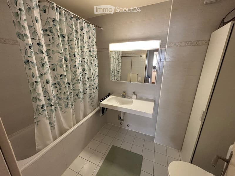 Bathroom with a bathtub, shower curtain, sink, mirror, and toilet. The walls are tiled, and a green bath mat is placed on the floor.