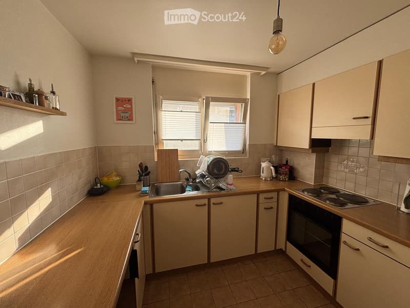 The kitchen features light wooden cabinets, a tiled backsplash, and a countertop with a sink, oven, and various kitchen items. There is a window above the sink, and the space is well-lit with natural light.