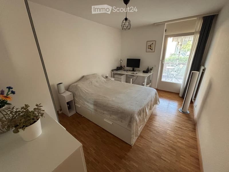 Cozy bedroom with wooden flooring, a large bed, a desk with a computer, a nightstand, and a balcony access.
