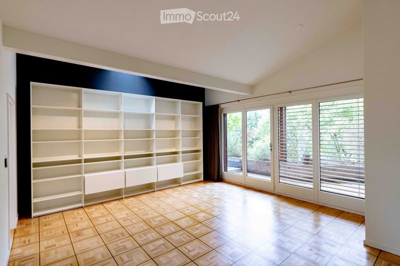 Empty room with wooden herringbone flooring, white built-in shelving unit, large windows with blinds, and a view of a garden area.