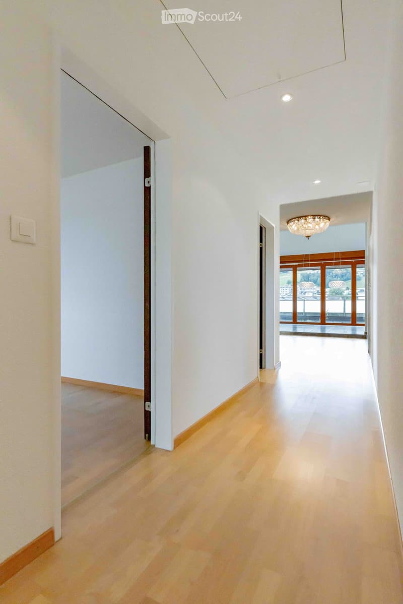 Empty hallway with light wooden flooring, white walls, and recessed ceiling lighting. Features a chandelier in the background and a view of a bright room beyond.
