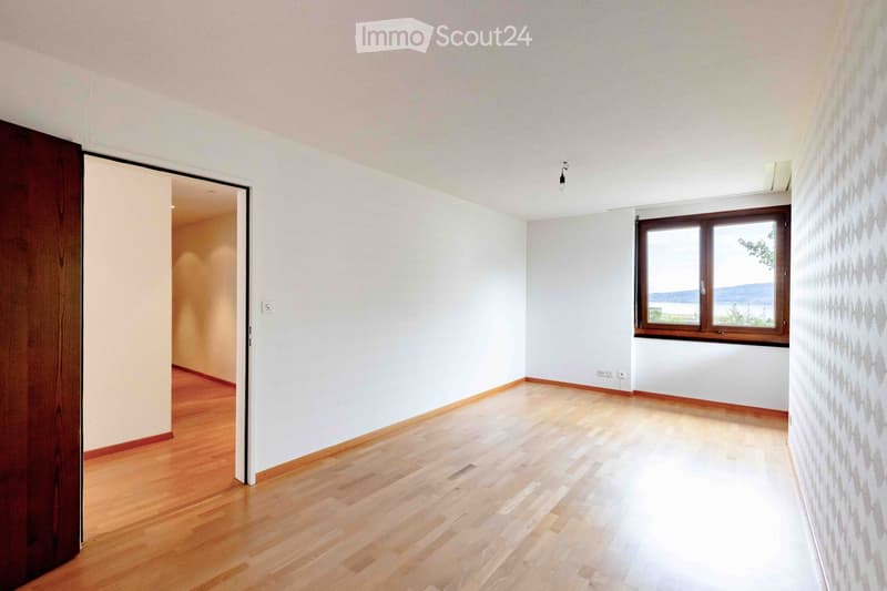 Empty room with wooden flooring, white walls, and a large window offering a scenic view. The room features a wooden door on the left and a light switch on the wall.