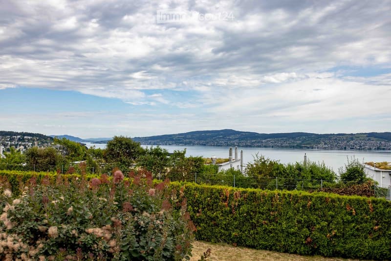 A well-maintained garden with neatly trimmed hedges and flowering shrubs, overlooking a body of water and distant cityscape.
