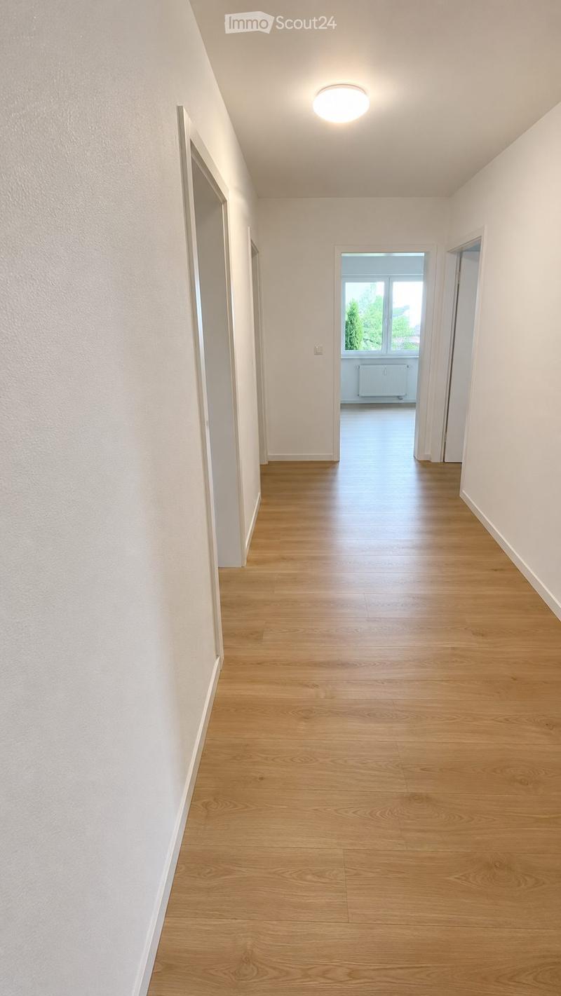 Empty hallway with light wooden flooring, white walls, and a ceiling light fixture. Two open doors lead to adjacent rooms, one with visible windows and a radiator.