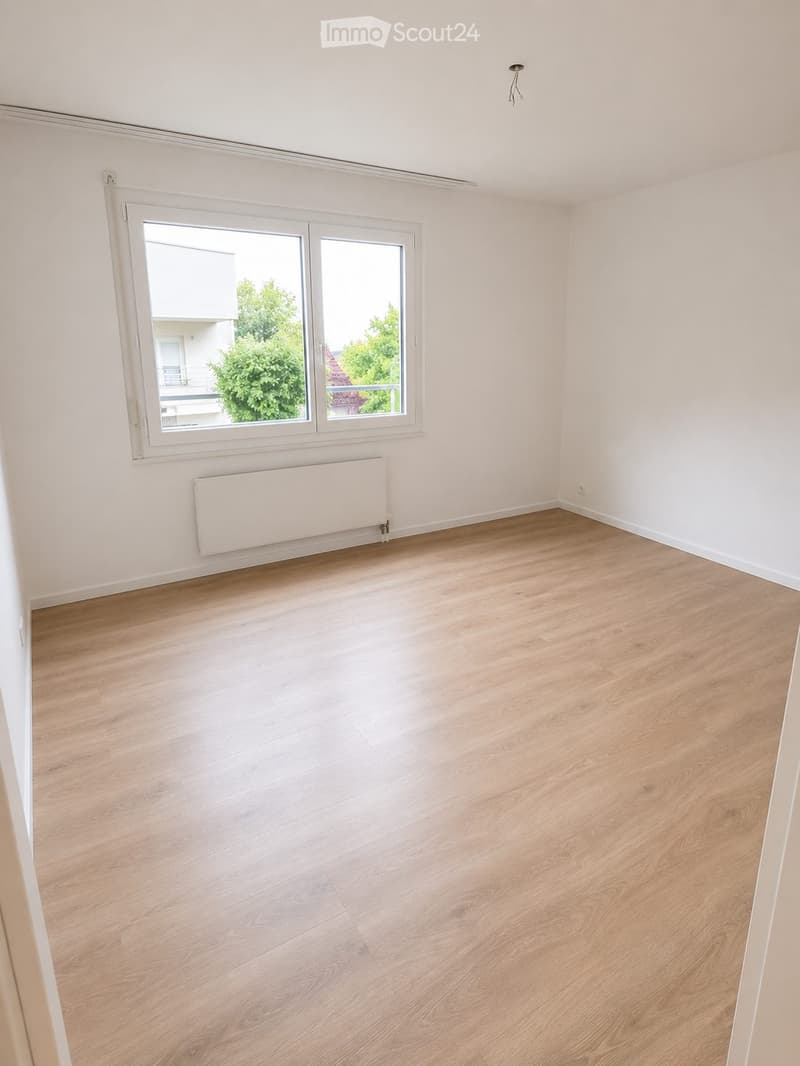 Empty room with light wooden flooring, white walls, and a large window with a view of greenery outside. A radiator is mounted below the window.