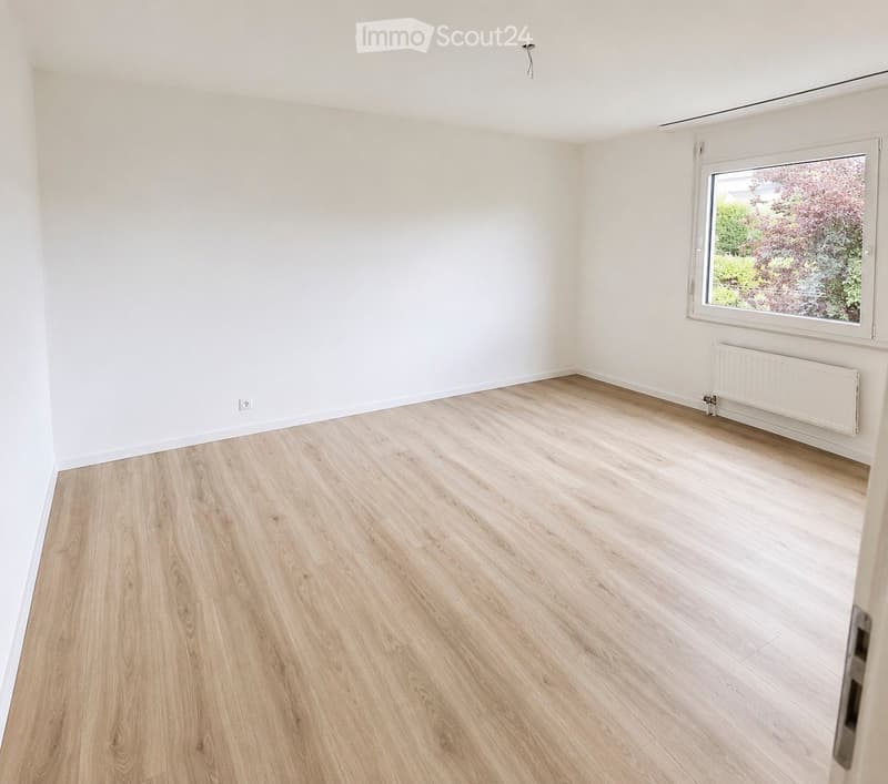 Empty room with light wooden flooring, white walls, and a large window with a view of greenery.
