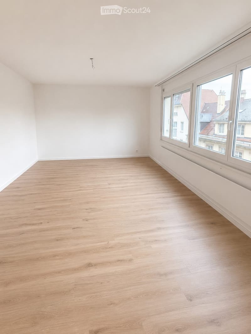 Empty room with light wooden flooring and white walls, large windows showing a view of rooftops, no furniture or equipment present.