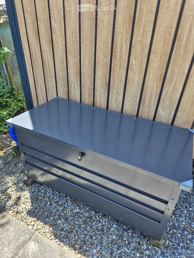 A sleek, modern outdoor bench with a dark grey metal frame and a flat, glossy top, positioned on a gravel surface next to a wooden fence.