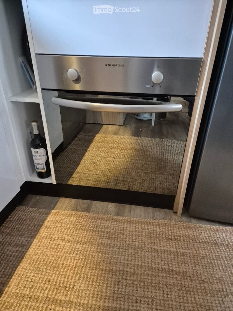A modern kitchen with a stainless steel oven, a small shelf holding a bottle, and a beige rug on the floor.