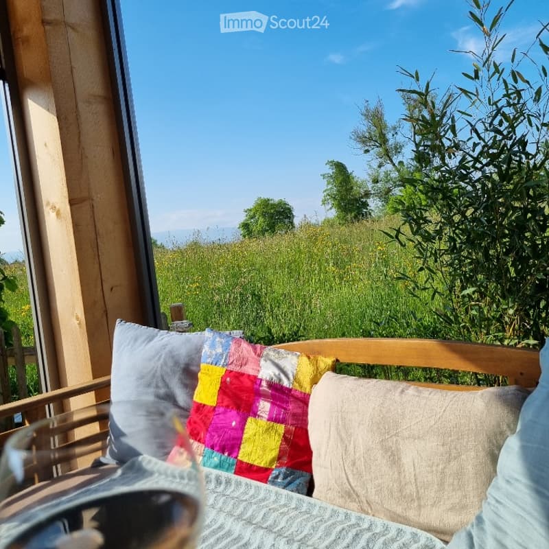 Balcony with wooden furniture, colorful patchwork quilt pillow, and a scenic view of a grassy field and trees under a clear blue sky.