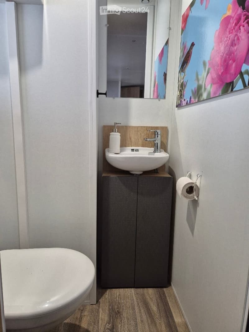 Modern bathroom with a white sink, dark grey cabinet, toilet, and a large mirror. The floor is wooden, and a picture frame is on the wall.
