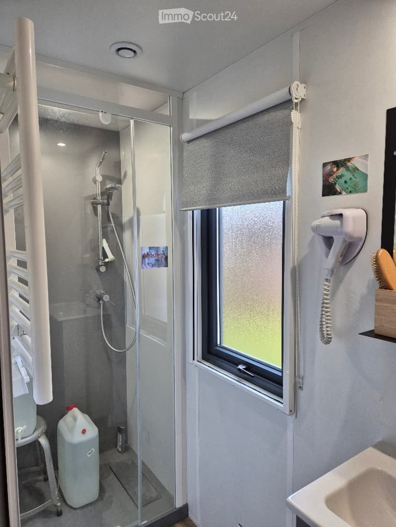 Modern bathroom with a glass-enclosed shower, a sink, a window with blinds, and a small stool holding a water bottle.