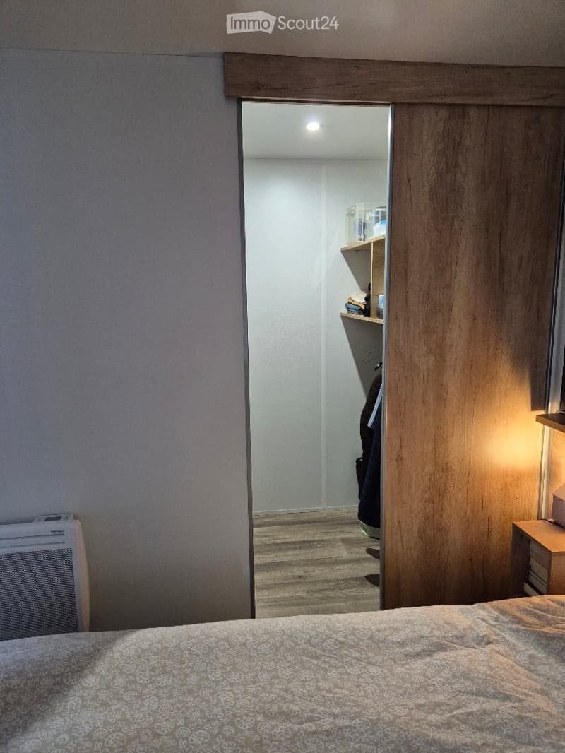 A modern bedroom with a neatly made bed, a nightstand with a lamp, a built-in wardrobe, and an open closet with shelves and hanging clothes.