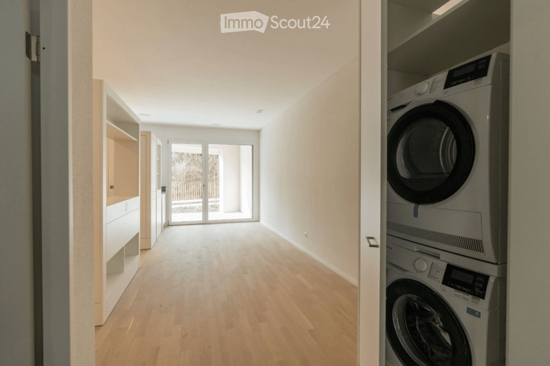 Empty hallway with wooden flooring, white walls, and a large glass door leading to an outdoor area. Laundry area visible with stacked washer and dryer.