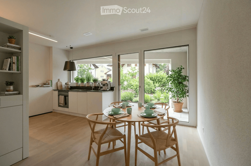 Modern kitchen with light-colored cabinets, a round dining table with four chairs, and a view of a garden through large windows.