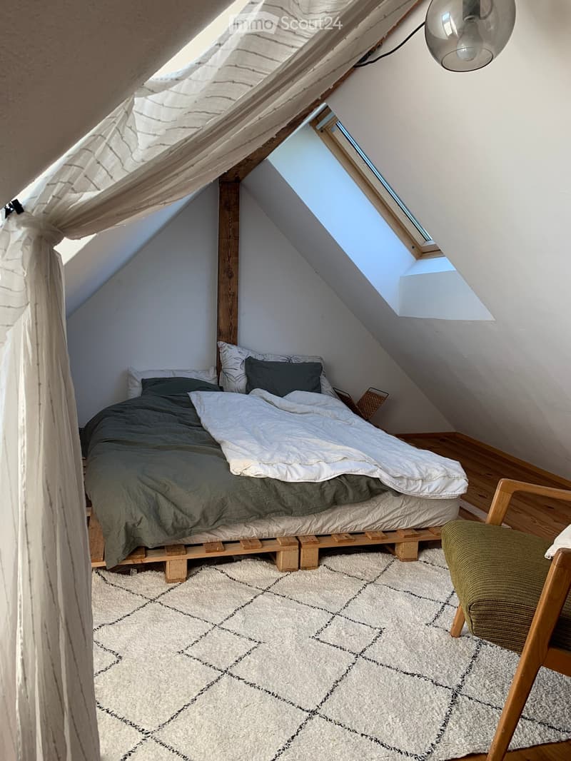 Cozy attic bedroom with a wooden bed, green bedding, a white shaggy rug, and a wooden chair near the window.