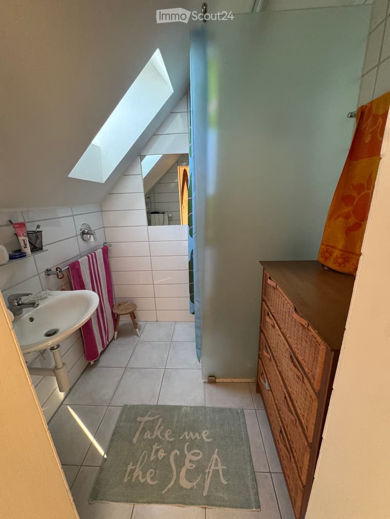 Cozy attic bathroom with a skylight, white tiles, a sink, a small wooden stool, a towel, a wooden cabinet, and a decorative rug that reads "Take me to the Sea".