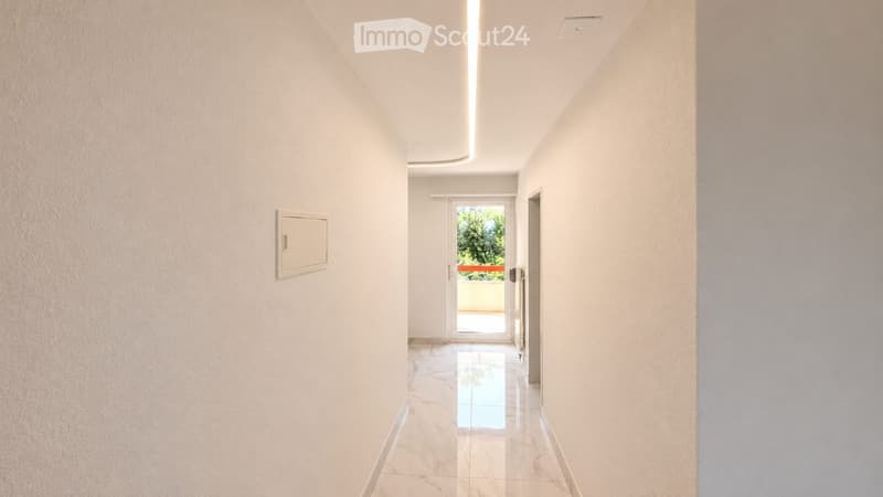 A clean, modern hallway with white walls, a polished marble floor, and a recessed ceiling light. A door at the end leads to a balcony or outdoor area.