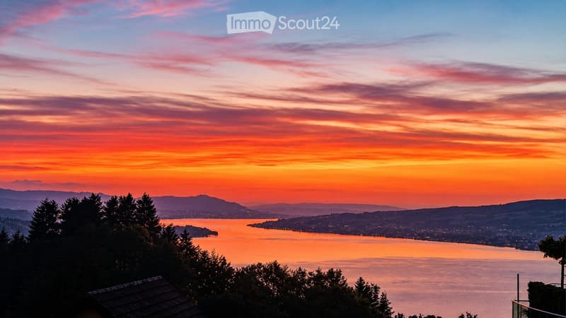Panoramic view of a lake at sunset, with city lights visible across the water and distant mountains in the background.