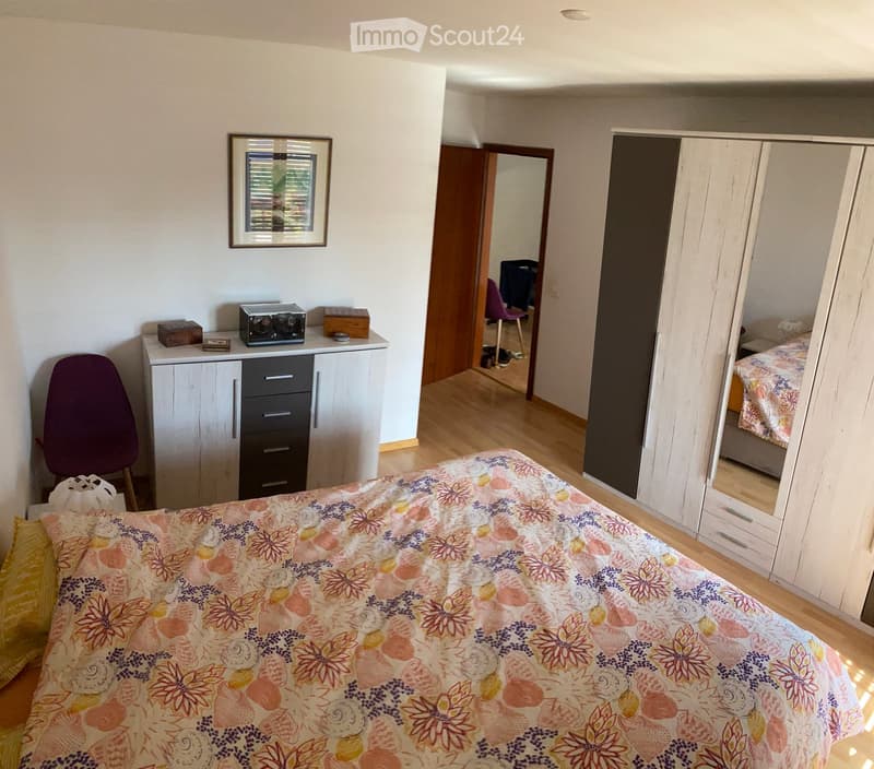 A well-lit bedroom with a bed covered in a floral-patterned duvet, a wooden wardrobe with mirrored doors, a small white cabinet with drawers, a purple chair, and a framed picture on the wall.