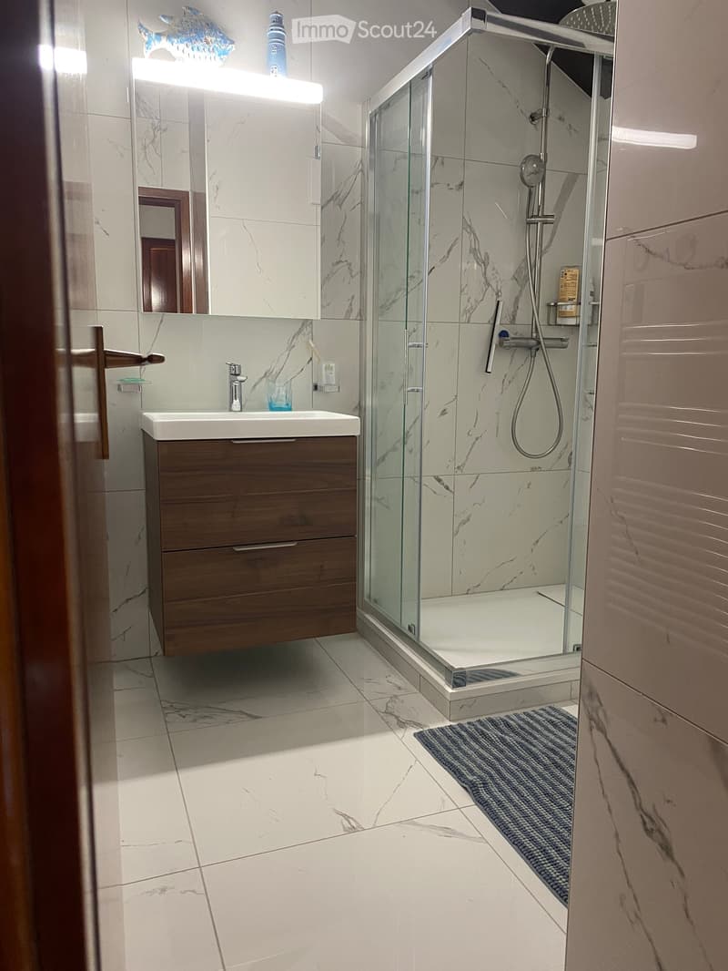 Modern bathroom with marble tiles, a wooden vanity with a sink, a glass-enclosed shower, and a mirror with built-in lighting.