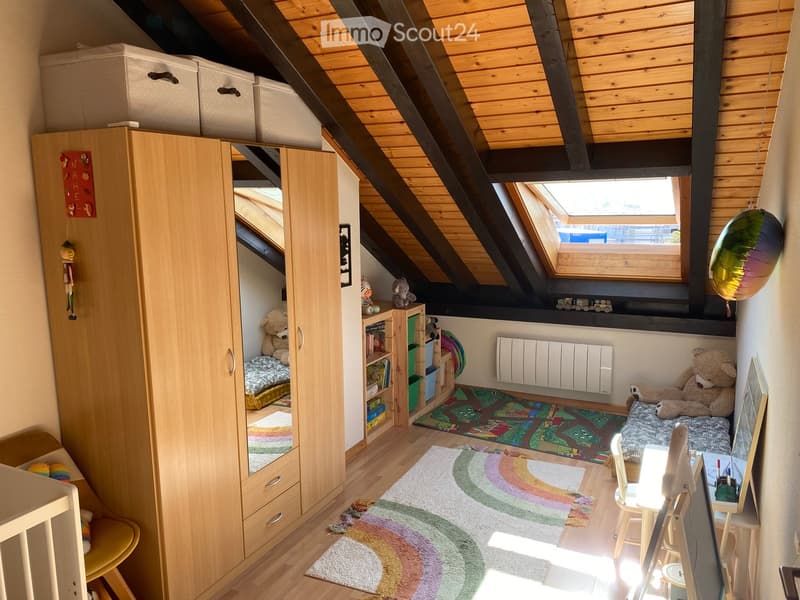 Cozy attic bedroom with wooden beams, skylight window, colorful rugs, and a variety of furniture including a wardrobe, bed, chair, table, and shelves.