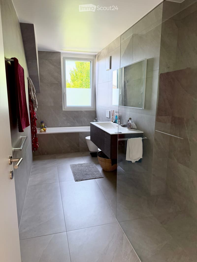 Modern bathroom with grey tiles, a bathtub, a sink with a mirror, a toilet, and a glass-enclosed shower area. Includes towels, a basket, and a small rug.