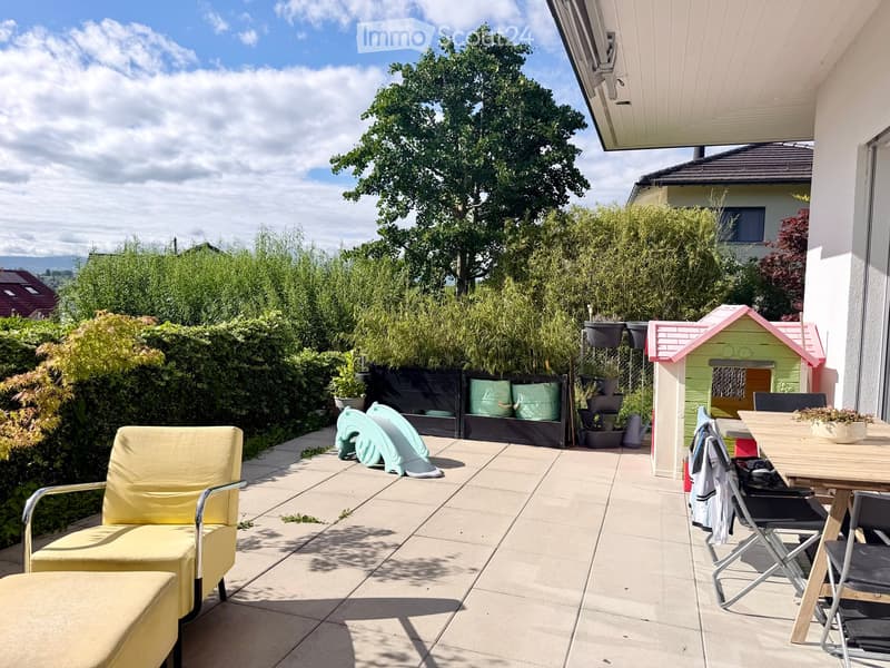 A sunny patio with a yellow chair, a small wooden table with chairs, a colorful toy house, and several potted plants. The area is surrounded by greenery and has a view of the landscape.