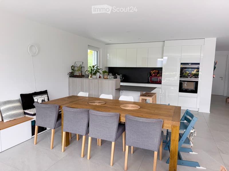 Modern kitchen and dining area with a wooden dining table, grey upholstered chairs, a built-in bench, white cabinets, and various appliances including an oven and microwave.