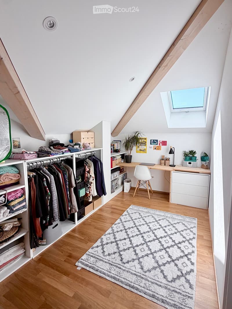 A cozy attic room with wooden flooring, a large window, a desk with a chair, a clothing rack, and a patterned rug.