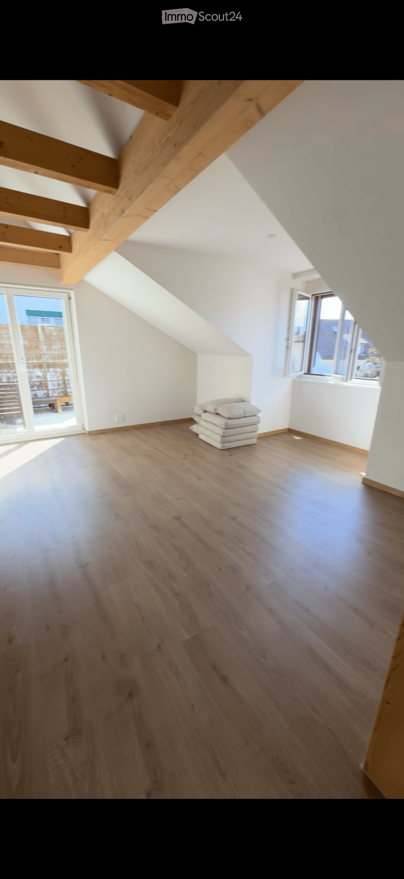 Empty attic room with wooden floor, white walls, exposed wooden beams, and large windows letting in natural light.