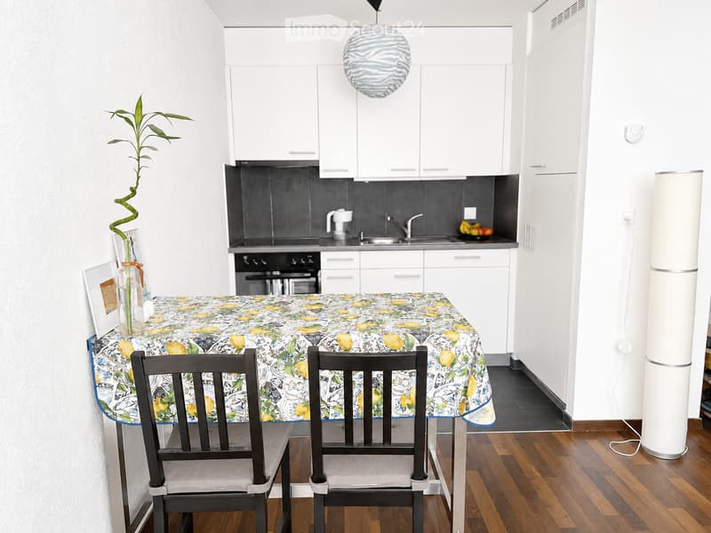 Modern kitchen with white cabinets, a round dining table with lemon-patterned tablecloth, two chairs, and a hanging lamp. The kitchen features a black backsplash, stainless steel appliances, and a small potted plant on the table.