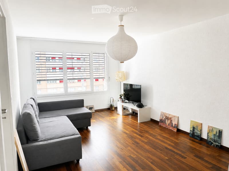 Modern living room with wooden flooring, large windows, grey sectional sofa, coffee table, TV, and decorative artwork.