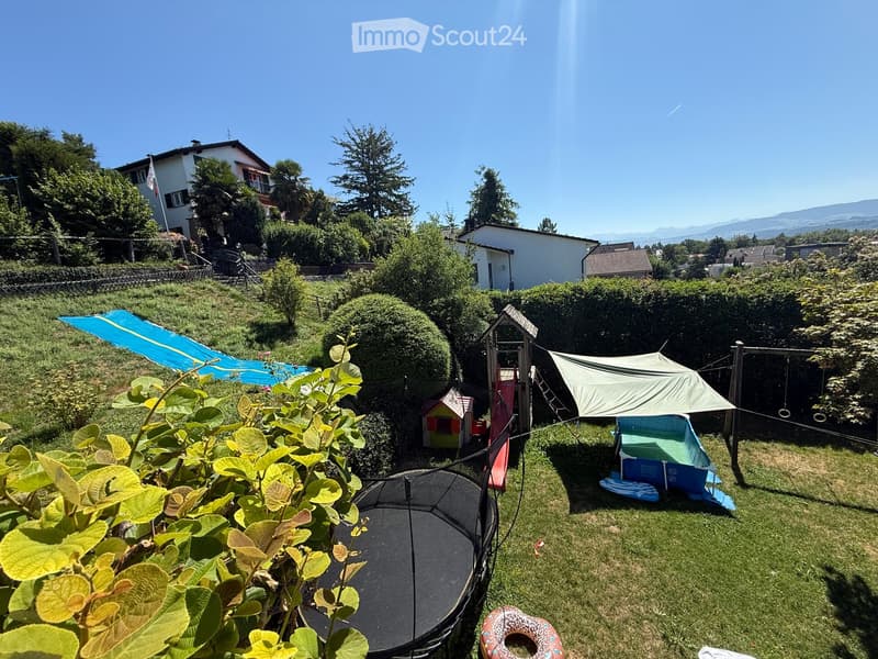 A well-maintained garden with a trampoline, slide, playhouse, and a small tent, surrounded by greenery and trees, with a view of nearby houses and mountains.