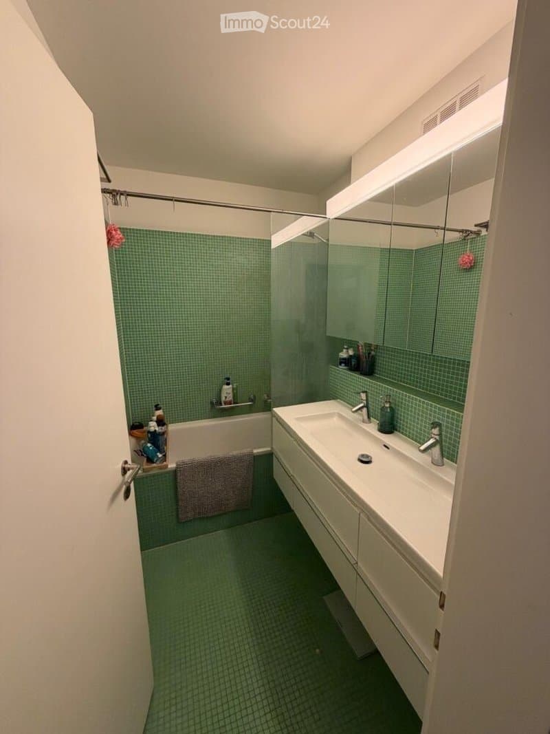Modern bathroom with green tiled walls and floor, featuring a bathtub, a double sink with a large mirror, and a glass-enclosed shower stall.