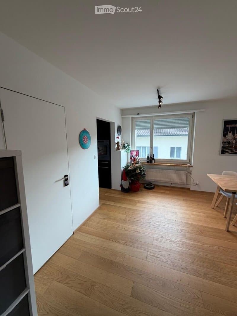 Empty room with wooden flooring, white walls, and a large window with blinds. A small Christmas tree is placed near the window, along with a few bottles and a radiator. A dining table and chairs are visible in the corner.