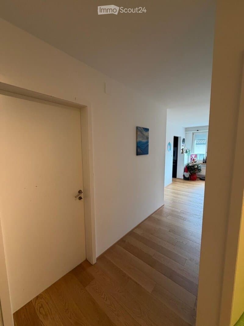 A narrow hallway with wooden flooring and white walls, leading to an adjacent room with a small Christmas tree and artwork on the walls.