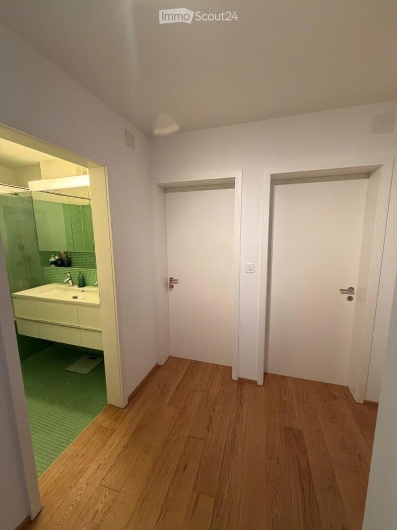 Empty room with wooden flooring and white walls, featuring two closed doors and a view into an adjacent bathroom with a sink, mirror, and green tiled shower area.
