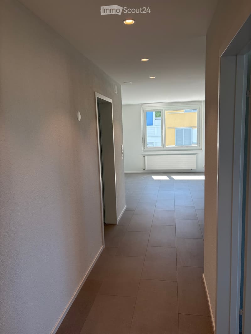 Empty hallway with tiled floor, white walls, recessed lighting, and a window at the end showing a view of a building.