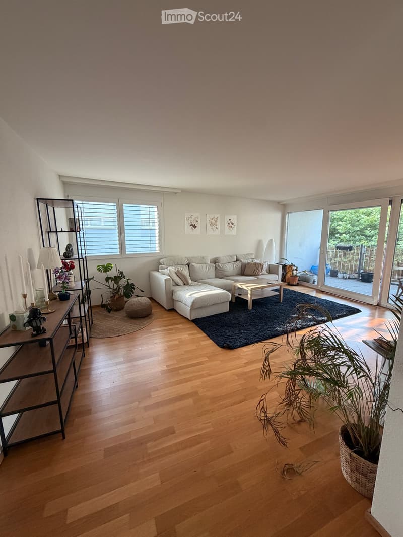 Modern living room with light-colored sectional sofa, coffee table, floor lamp, side table, and potted plants. Large windows and sliding glass doors lead to a patio area.
