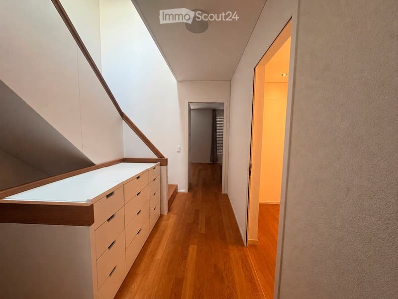 Modern hallway with wooden flooring, white walls, built-in storage unit with drawers, and a skylight providing natural light. The hallway leads to another room with a visible door and window.