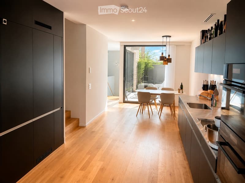 Modern kitchen with light wood flooring, grey cabinets, stainless steel appliances, and a dining area with a table and chairs. The space is well-lit with natural light from a large window and pendant lights.