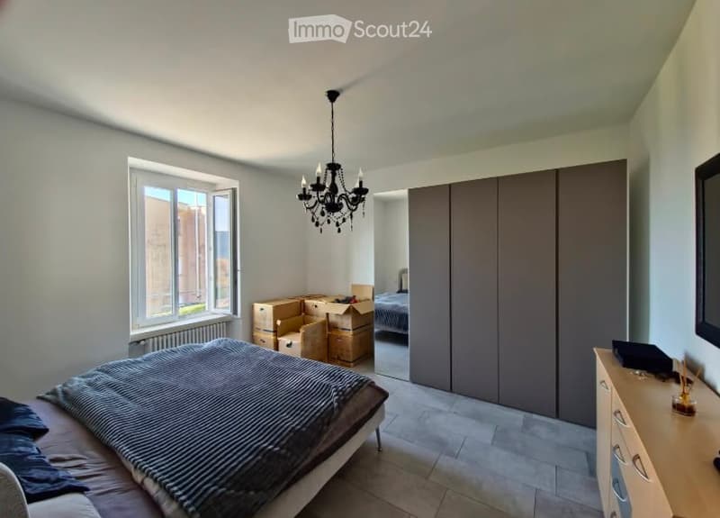 Modern bedroom with a large window, a bed with striped bedding, a wardrobe, a dresser, and a chandelier. Some boxes are visible, indicating the room is being unpacked or organized.