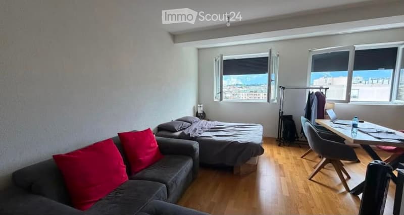 A compact studio with a sofa, bed, small table, and chair, featuring large windows with a city view.