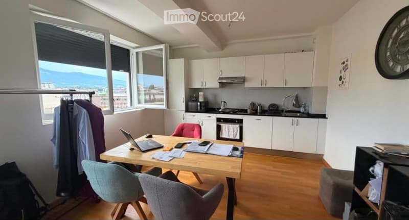 A modern, well-lit room with a wooden table, laptop, and chairs. The space includes a small kitchen area with white cabinets, a sink, and various appliances.