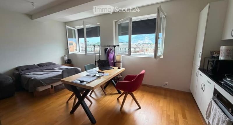 A modern, well-lit bedroom with a large window offering a scenic view. The room features a bed with dark bedding, a wooden desk with a laptop, a pink chair, and a small kitchen area with cabinets and a microwave.