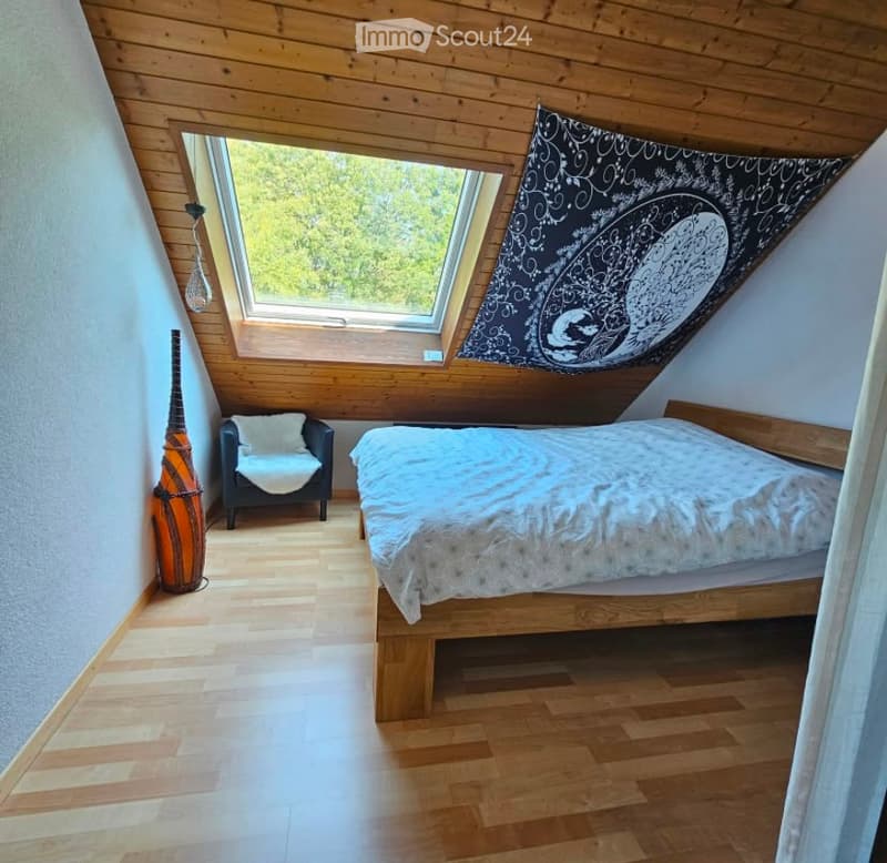 Cozy attic bedroom with wooden slanted ceilings, skylight windows, a bed, a small chair, a decorative vase, and a wall hanging.
