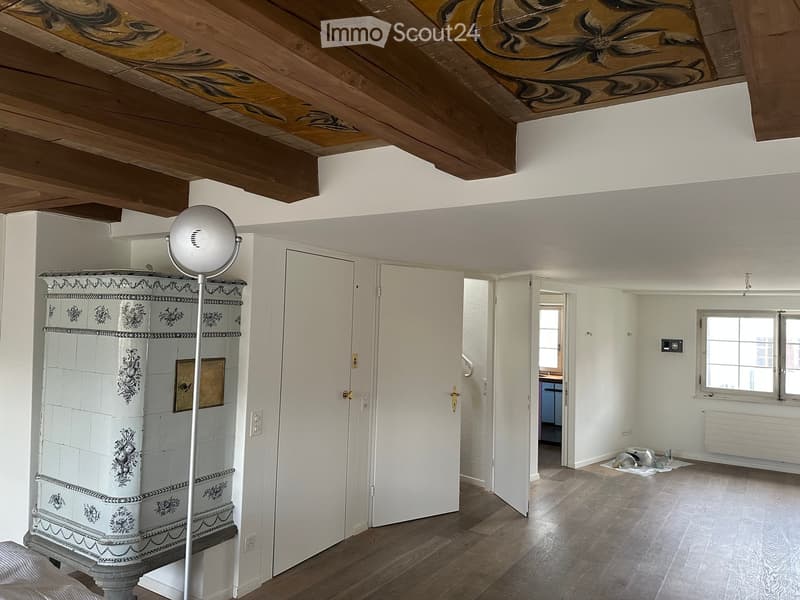 An unfurnished room with wooden flooring, white walls, and a decorative cabinet. The room features a large window, a door leading to another space, and a modern spherical lamp on a stand.